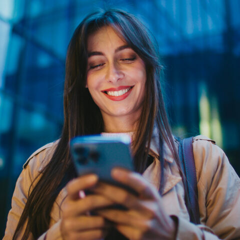 Woman smiling while looking at her smartphone in a city setting with blue evening light.